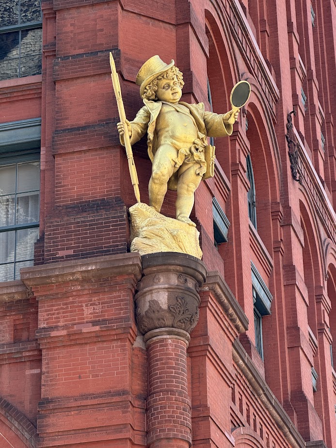 The gilded Puck statue on the Puck Building at 295 Lafayette Street — top-hatted, holding a quill and a raised hand mirror.