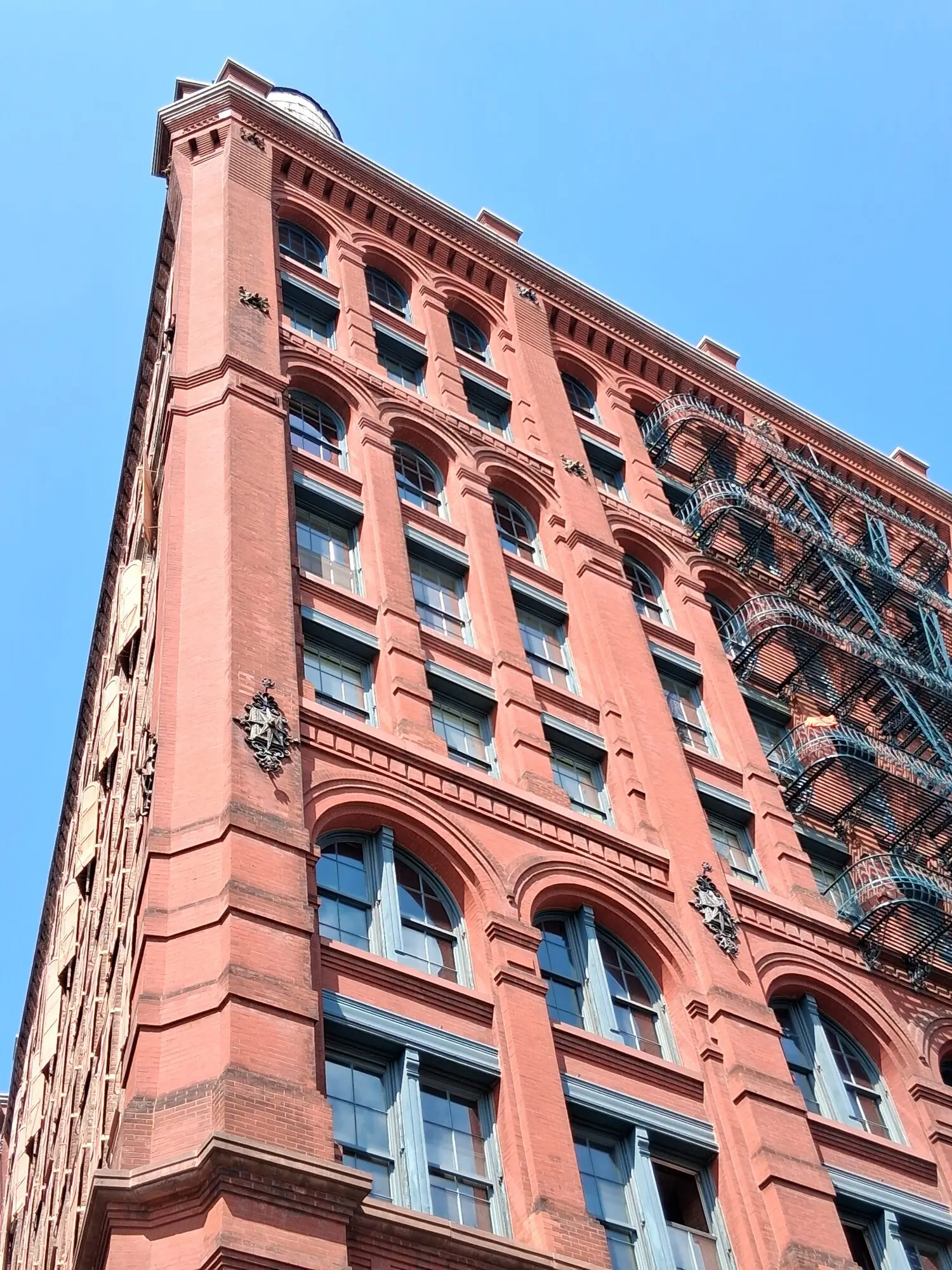 Looking straight up the brick corner of the Puck Building against the sky — the gilded figure high on the upper floor, the street far below, no one looking up.