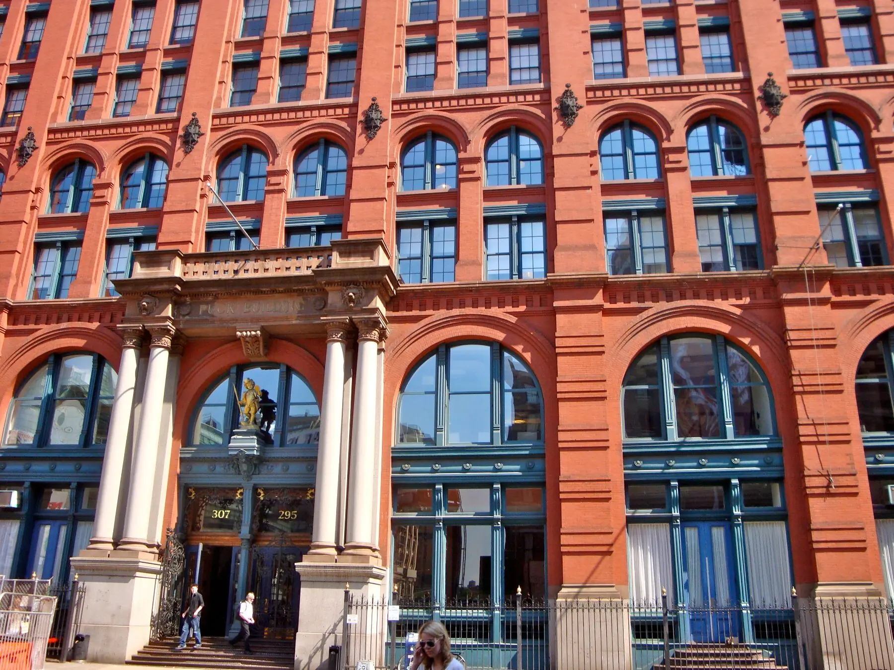 The gilded statue of Puck on the corner of the Puck Building — a golden cherub in a top hat and open frock coat against red brick, holding a tall pen in one hand and raising a hand mirror with the other.