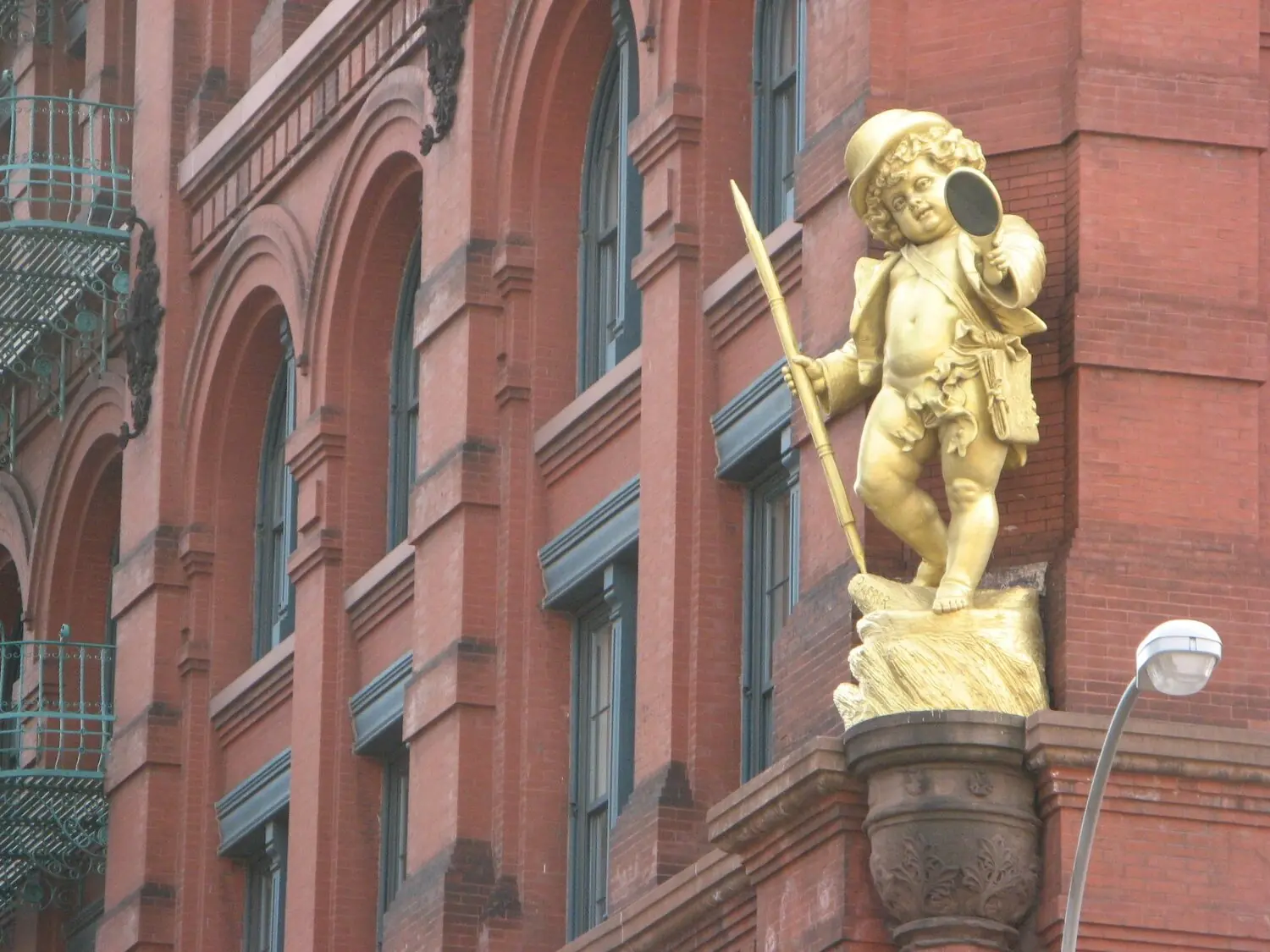 The gilded Puck against the red brick of the building, top hat and open coat, a tall pen held like a staff in one hand and a hand mirror raised in the other.