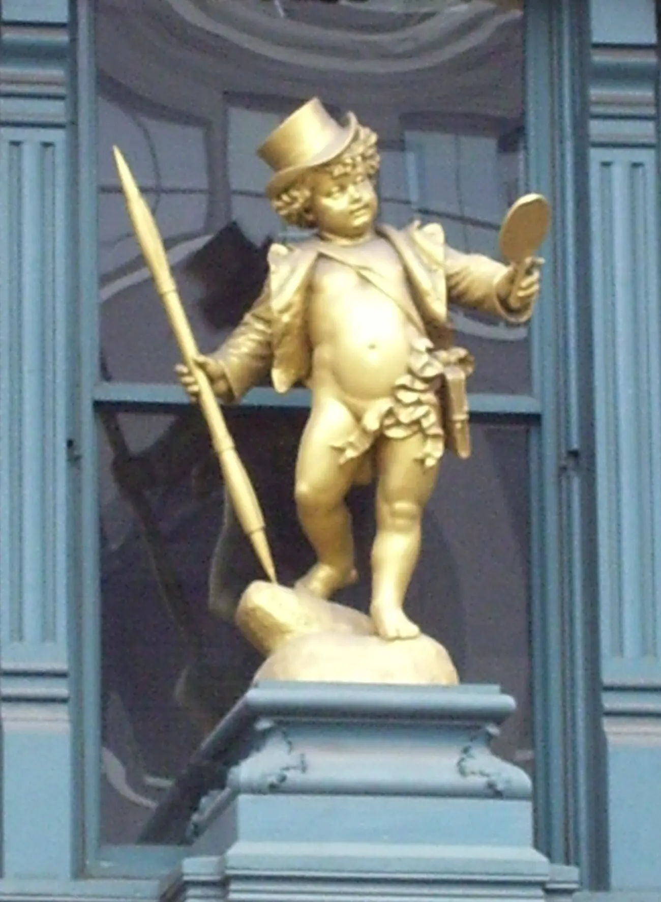 Close view of the gilded Puck on his pedestal — a cherub in a top hat and open frock coat, holding a tall pen in one hand and raising a hand mirror in the other.